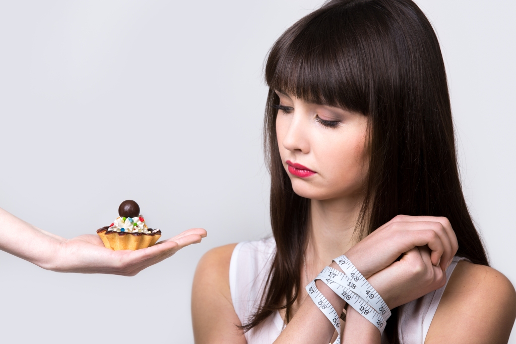 Living well in the 21st century - Limassol, Cyprus. Young dieting woman sitting in front of delicious cream tart cake with hands tied with measuring tape, looking at forbidden food with longing and hungry expression, studio, gray background, isolated.