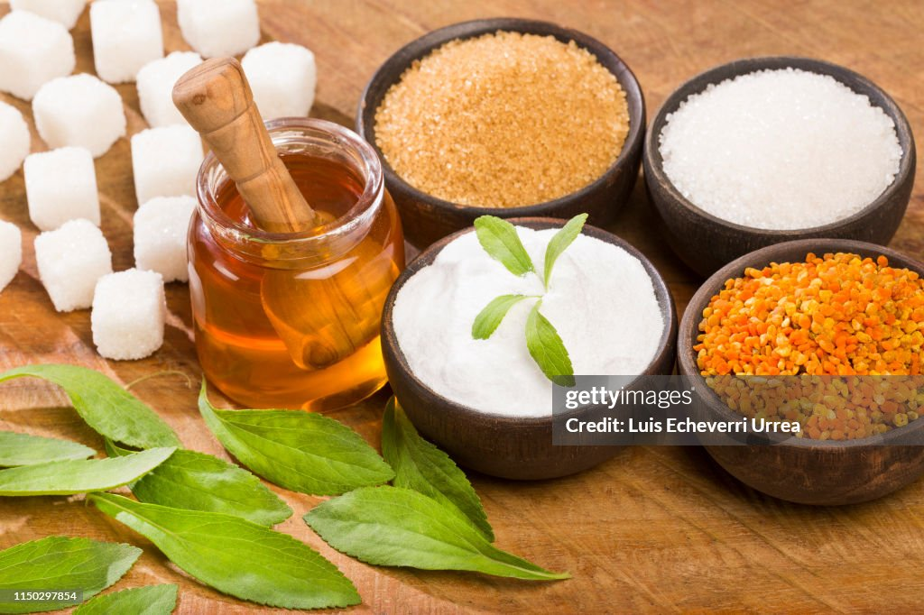 Living well in the 21st century-Limassol, Cyprus. A picture of sugars-the picture above shows a variety of sugars from honey, stevia plant, bee pollen, white sugar cubes, white, and brown sugar on a brown background. The honey container holds a small brown spoon, and several green stevia plant stems are laid out on the table. The sugars are places in four small black bowls, with a stevia stem placed in the middle of the stevia sugar bowl.  